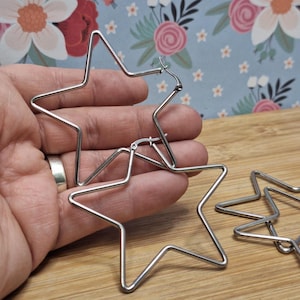 May include: Silver-coloured star-shaped hoop earrings. The earrings are made of thin metal and have a simple, geometric design. The earrings are shown against a floral background.