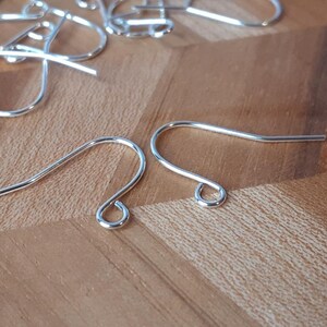 May include: Two silver earring hooks, also known as ear wires, are shown on a wooden surface. The hooks are made of metal and have a simple, classic design.
