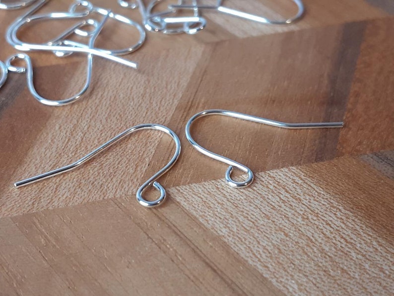 May include: Two silver earring hooks, also known as ear wires, are shown on a wooden surface. The hooks are made of metal and have a simple, classic design.