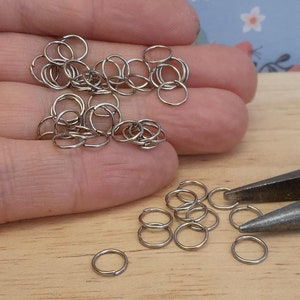 May include: A close-up of a pile of silver jump rings, some of which are scattered on a wooden surface. There are also a pair of pliers in the image.