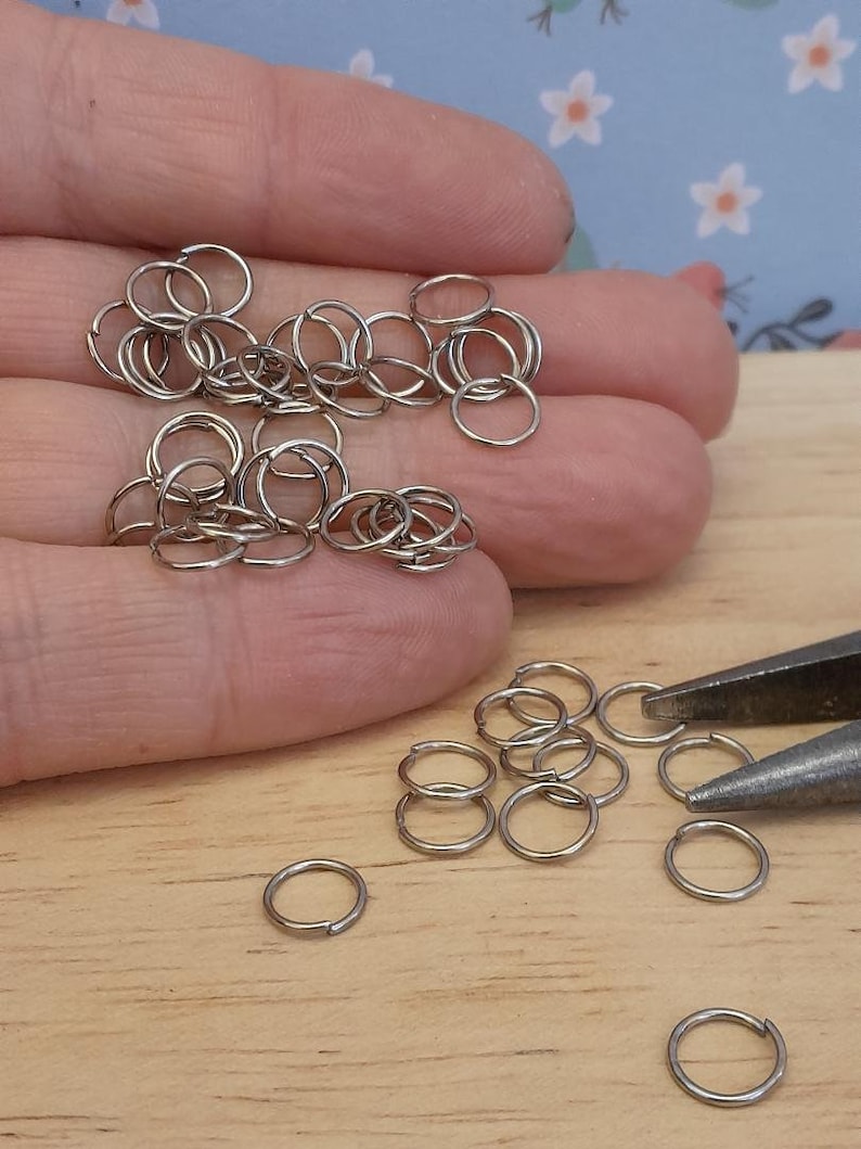 May include: A close-up of a pile of silver jump rings, some of which are scattered on a wooden surface. There are also a pair of pliers in the image.