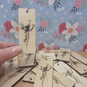 May include: Wooden poppy flower bookmarks with a small metal ring at the top. The poppy flower and the word "Poppy" are etched into the wood. The background is a floral pattern with pink and white flowers.