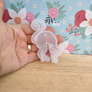May include: A clear silicone mould shaped like a cat with a curled tail. The mould is held in a hand against a floral background.