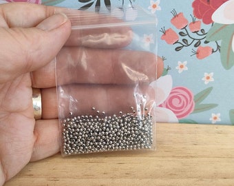 May include: A clear plastic bag filled with small, silver metal beads. The bag is held in a hand.