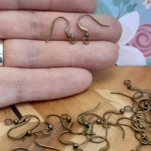 Może przedstawiać: Zbliżenie na stos 20 złotych haczyk&oacute;w do kolczyk&oacute;w. Haczyki są wykonane z metalu i mają małą kulkę na g&oacute;rze. Haczyki są ułożone w przypadkowy wz&oacute;r na drewnianej powierzchni.