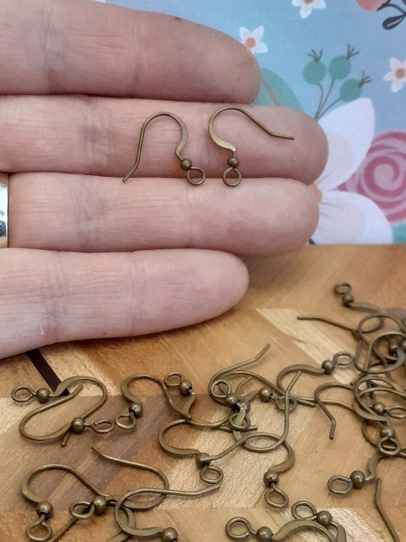 Może przedstawiać: Zbliżenie na stos 20 złotych haczyk&oacute;w do kolczyk&oacute;w. Haczyki są wykonane z metalu i mają małą kulkę na g&oacute;rze. Haczyki są ułożone w przypadkowy wz&oacute;r na drewnianej powierzchni.