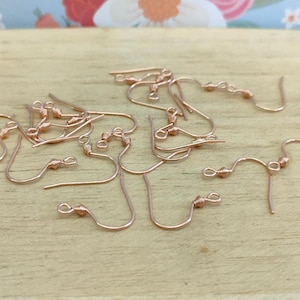 May include: A close-up of a group of rose gold earring hooks. The hooks are made of metal and have a small loop at the top. The hooks are arranged in a random pattern on a wooden surface.