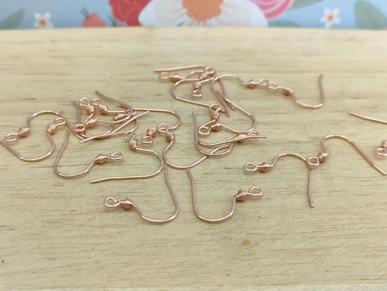May include: A close-up of a group of rose gold earring hooks. The hooks are made of metal and have a small loop at the top. The hooks are arranged in a random pattern on a wooden surface.