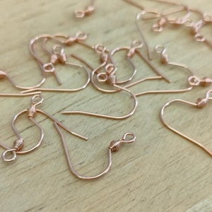 May include: A close-up of a group of rose gold earring hooks. The hooks are made of metal and have a small, twisted loop at the top.
