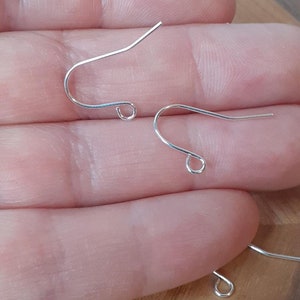 May include: Three silver earring hooks, also known as ear wires, are shown on a hand. The hooks are made of metal and have a simple design.