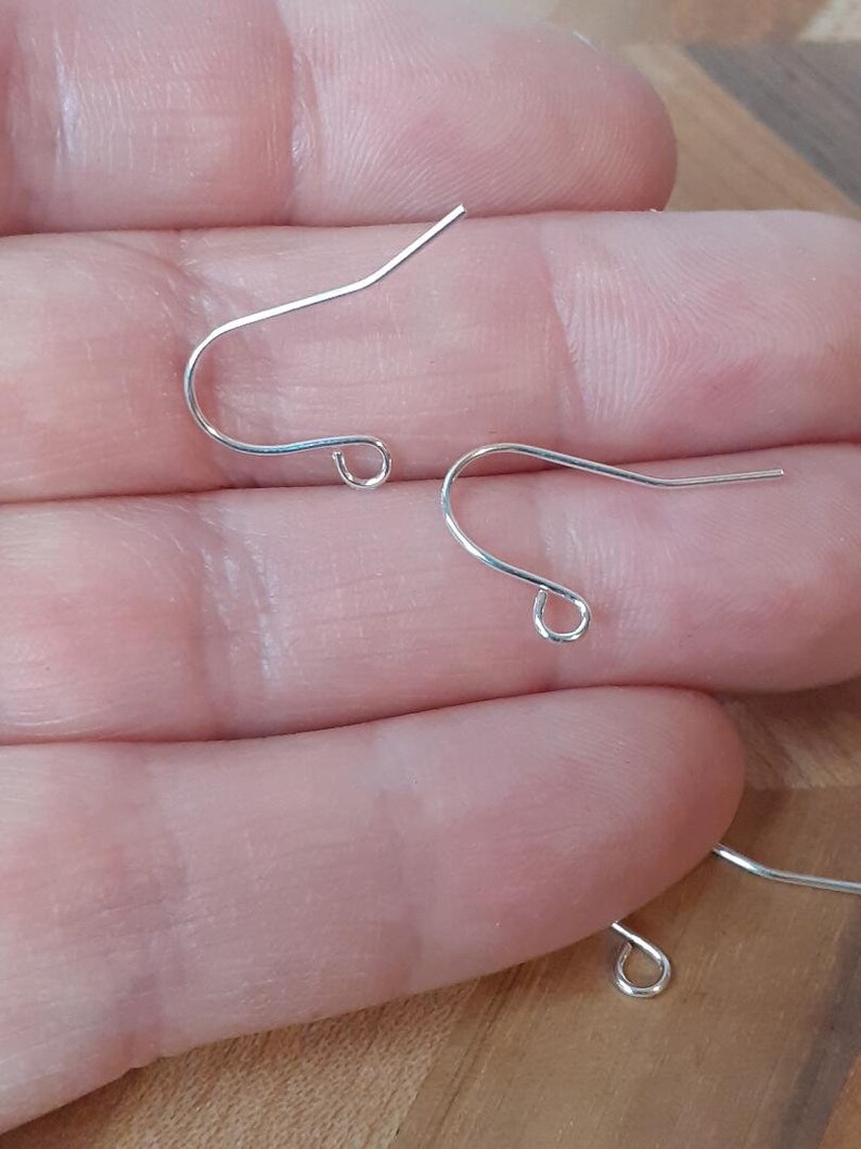 May include: Three silver earring hooks, also known as ear wires, are shown on a hand. The hooks are made of metal and have a simple design.