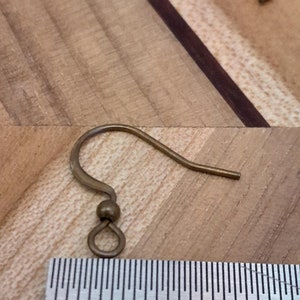 Może przedstawiać: Złoty haczyk do kolczyk&oacute;w z małą pętelką na dole. Haczyk jest mierzony linijką, pokazując, że ma nieco ponad 2,5 cm długości.