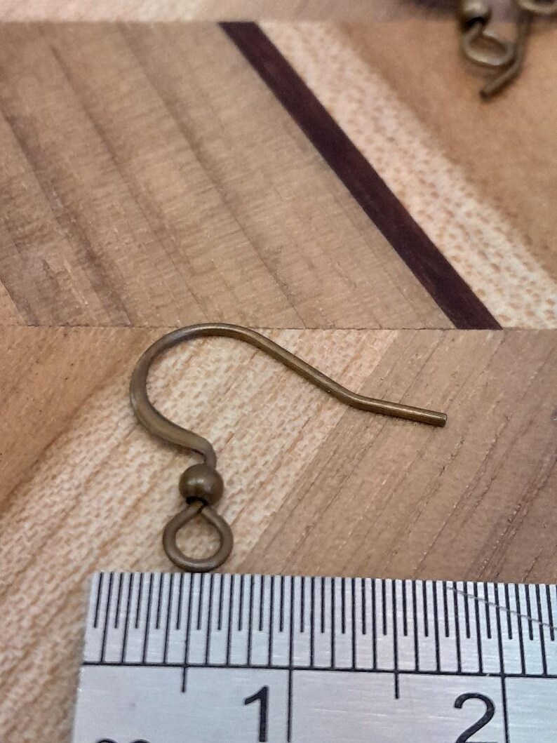 Może przedstawiać: Złoty haczyk do kolczyk&oacute;w z małą pętelką na dole. Haczyk jest mierzony linijką, pokazując, że ma nieco ponad 2,5 cm długości.