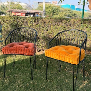 May include: Two black metal outdoor chairs with orange and yellow cushions sit on a grassy lawn. A potted plant with large green leaves and a gold-colored pot is to the left. The chairs have a curved back design.