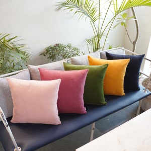 May include: Six decorative throw pillows in various colors, including pink, red, green, yellow, and navy blue, arranged on a dark blue couch. The pillows are made of a soft, velvety fabric.