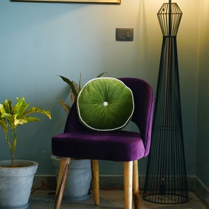 May include: A purple velvet chair with wooden legs and a round green cushion with white piping. A tall black metal floor lamp and a potted plant are in the background. A framed painting hangs on the wall.