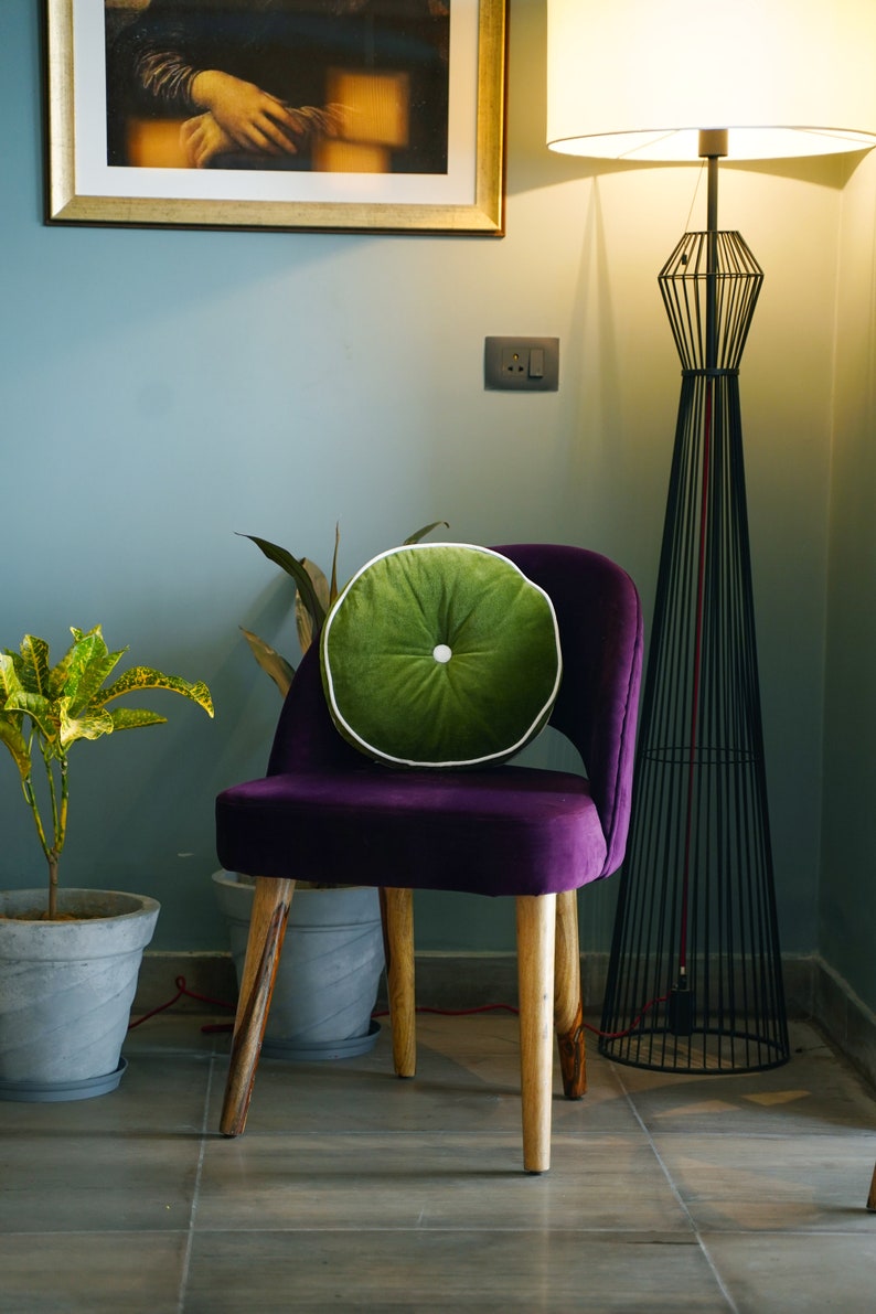 May include: A purple velvet chair with wooden legs and a round green cushion with white piping. A tall black metal floor lamp and a potted plant are in the background. A framed painting hangs on the wall.