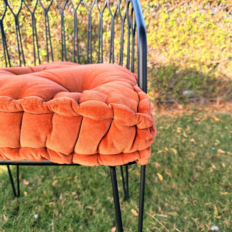 May include: Close-up of a chair with a tufted, orange cushion. The cushion is made of a soft, velvety material. The chair has a black metal frame with thin, hairpin legs and a decorative back.