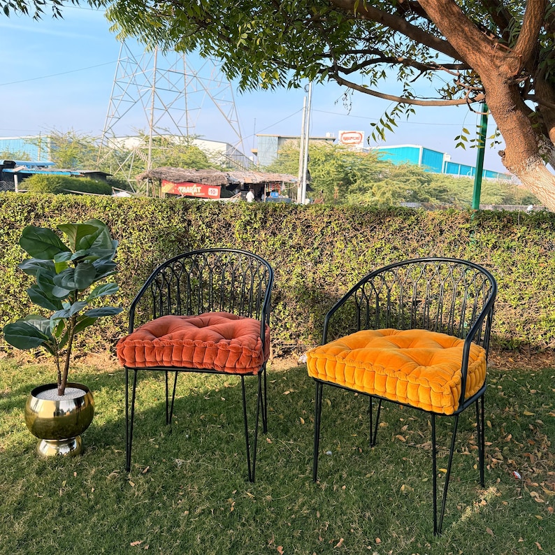 May include: Two black metal outdoor chairs with orange and yellow cushions sit on a grassy lawn. A potted plant with large green leaves and a gold-colored pot is to the left. The chairs have a curved back design.