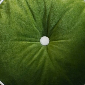 May include: A round, green velvet cushion with a white button in the center. The cushion has a tufted design, with the fabric radiating outwards from the button. The color is a vibrant, medium green.