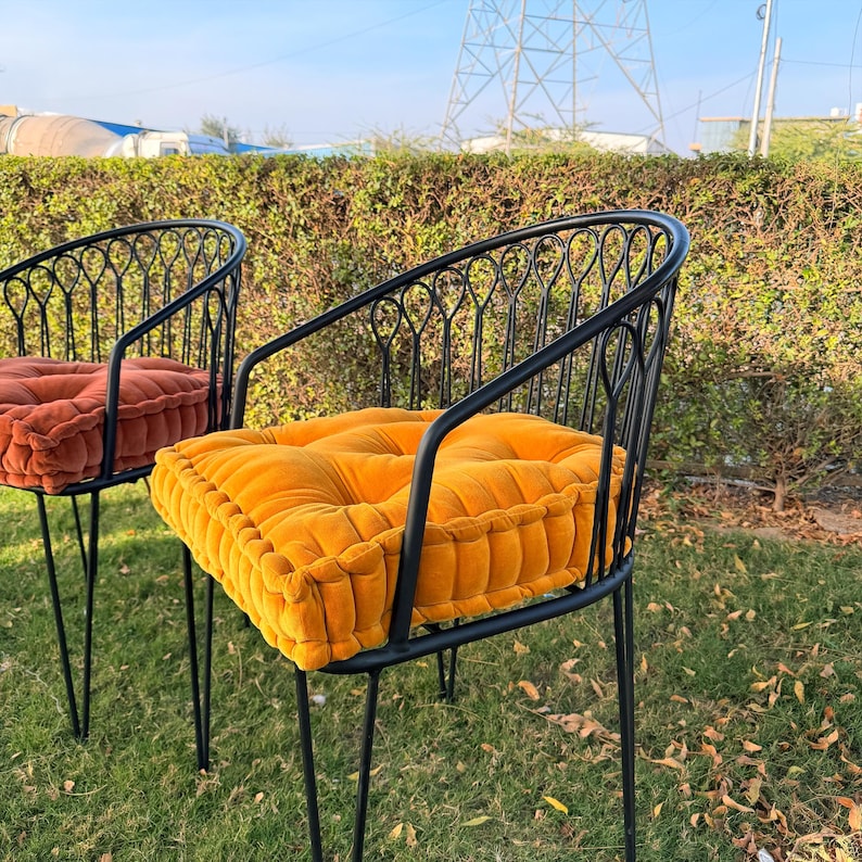 May include: Two black metal chairs with rounded backs and thin legs. Each chair has a square, tufted cushion. One cushion is orange, the other is yellow. The chairs are outdoors on grass, with a green hedge in the background.