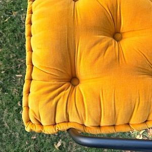 May include: Close-up of a tufted, square cushion in a vibrant mustard yellow color. The cushion has a plush, velvety texture with button detailing. The cushion is part of a chair, with a black metal frame visible at the bottom.