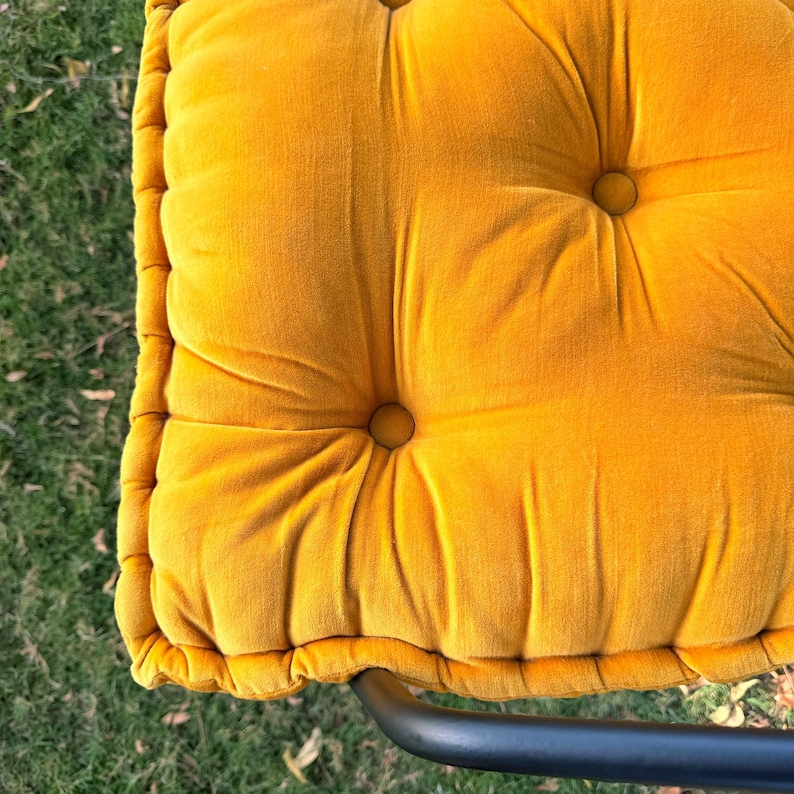 May include: Close-up of a tufted, square cushion in a vibrant mustard yellow color. The cushion has a plush, velvety texture with button detailing. The cushion is part of a chair, with a black metal frame visible at the bottom.