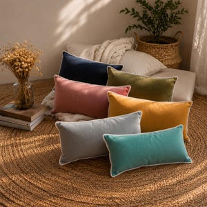 May include: A collection of rectangular throw pillows in various colors, including navy, pink, olive green, yellow, gray, and teal. Each pillow has a white trim and is arranged on a woven jute rug. The pillows are soft and plush.