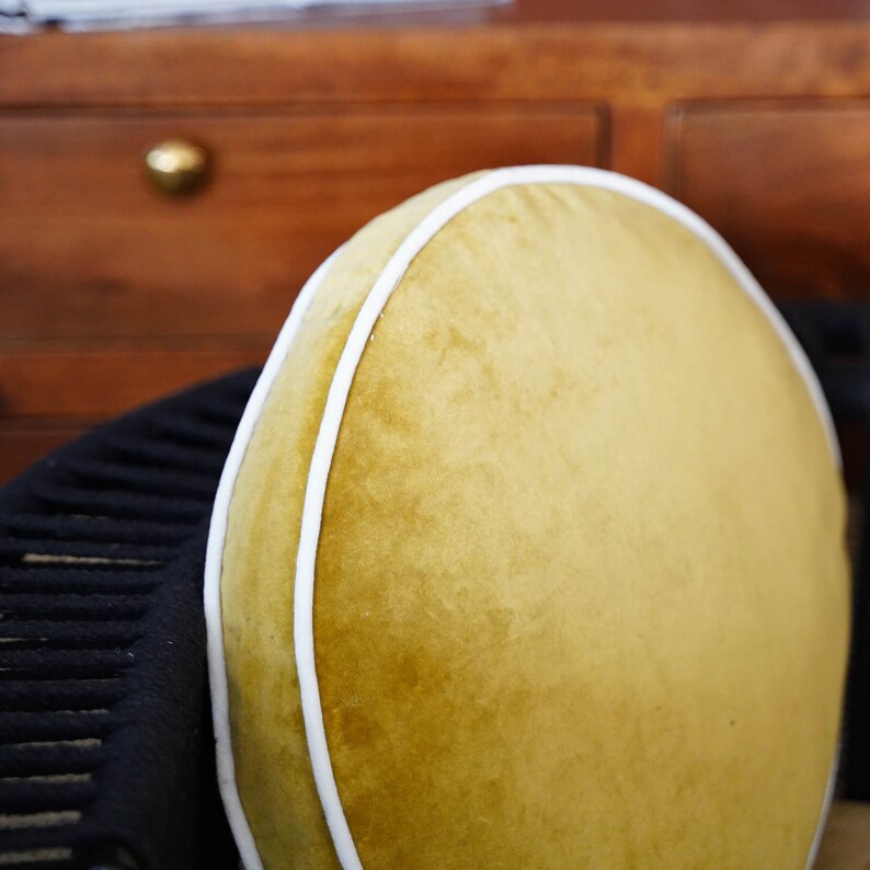 May include: A round, mustard-yellow velvet cushion with a white trim. The cushion is set against a dark background, with a glimpse of a wooden cabinet in the background.