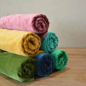 May include: A stack of rolled-up velvet fabric in various colors, including pink, burgundy, yellow, teal, blue, and green. The fabric is arranged on a wooden surface, with a neutral background.