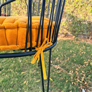 May include: A close-up of a black metal chair with a round seat and back. The seat cushion is a vibrant orange-yellow color, and the chair has a matching ribbon tied to the frame. The chair is outdoors.