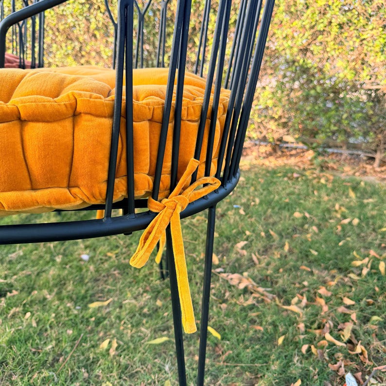 May include: A close-up of a black metal chair with a round seat and back. The seat cushion is a vibrant orange-yellow color, and the chair has a matching ribbon tied to the frame. The chair is outdoors.