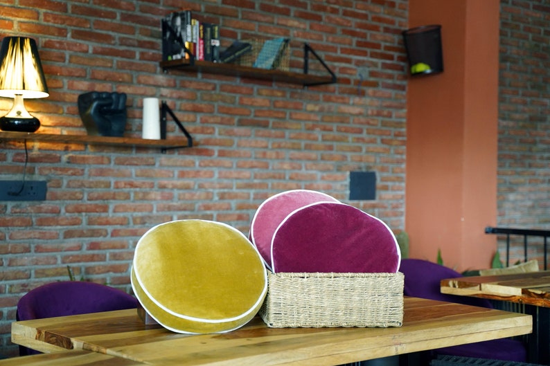 May include: A wooden table displays three velvet cushions in yellow, pink, and burgundy, each with a white trim. The cushions are arranged with a woven basket. The background features a brick wall and shelves.
