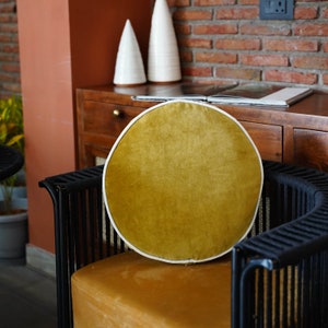 May include: A round, mustard-yellow velvet cushion with a white trim sits on a black chair. The chair has a woven design and a curved back. The background includes a wooden cabinet, decorative white cones, and a brick wall.