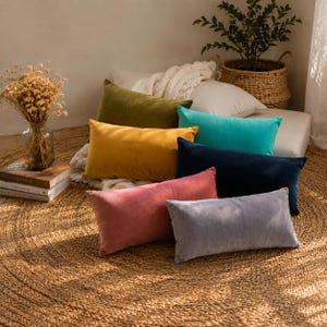 Puede incluir: Una colección de cojines rectangulares de terciopelo en varios colores, incluyendo verde oliva, amarillo mostaza, turquesa, azul marino, rosa empolvado y gris. Los cojines están colocados sobre una alfombra de yute tejida, creando un ambiente acogedor.