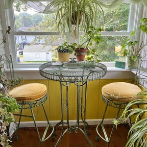 Puede incluir: Un juego de bistró vintage con una mesa redonda de metal verde y dos taburetes a juego. Los taburetes tienen cojines amarillos. El conjunto está rodeado de plantas en macetas y está colocado contra una pared amarilla y una ventana que da a un vecindario.
