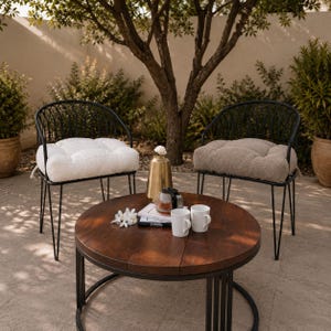 May include: Two black metal chairs with arched backs and tufted cushions, one white and one beige, positioned around a round dark wood coffee table. The table holds a gold vase, a glass carafe, two white mugs, and two books with visible text.