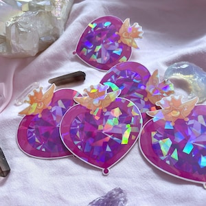 Könnte beinhalten: Herzförmige Aufkleber mit einem facettierten, irisierenden Design in Rosa- und Lilatönen. Jeder Aufkleber hat eine goldfarbene Krone und Flügel. Die Aufkleber sind auf einem rosa Stoff verteilt, mit Kristallen und Edelsteinen im Hintergrund.