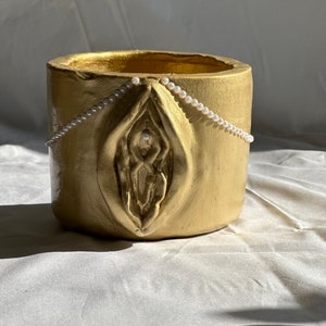 May include: A gold-colored, cylindrical ceramic planter with a sculpted vulva design on the front. A string of small, white pearls is draped across the top. The planter is set against a white, textured background.