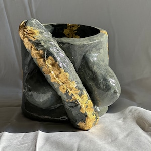 May include: A gray ceramic planter with a gold leaf design. The planter is shaped like a torso and has a rounded, organic form.