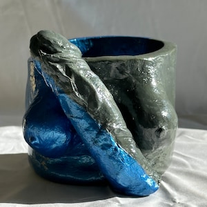 May include: A gray and blue ceramic planter shaped like a human figure. The planter has a textured surface and is designed to hold plants.