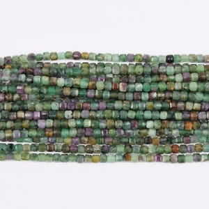 Peut inclure: Des brins de perles d'émeraude facettées de différentes nuances de vert, de violet et de marron. Les perles sont petites, de forme carrée et enfilées ensemble, créant une surface texturée. Les perles sont présentées sur un fond blanc.
