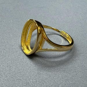 Puede incluir: Un anillo dorado con un engaste ovalado. La banda del anillo se divide en dos partes para sostener el engaste. El anillo está hecho de un metal brillante y reflectante.
