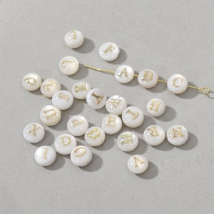 May include: White round beads with gold letters of the alphabet. The beads are scattered on a light grey surface. A gold wire with three beads on it is in the top right corner of the image. The beads on the wire spell out the letters "A B C".