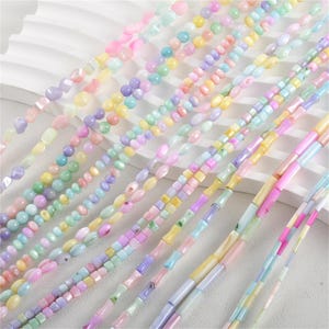 May include: Assortment of pastel colored beads made from natural materials. The beads are in various shapes and sizes, including round, oval, square, and rectangular. The beads are strung on a white background.
