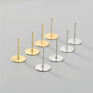 Könnte beinhalten: Acht Ohrringrohlinge, vier goldfarben und vier silberfarben, mit runden Basen und Stiften. Die Ohrringrohlinge sind in zwei Reihen auf einer weißen Oberfläche angeordnet.