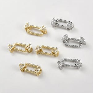 Pode incluir: Seis pingentes conectores para fazer joias. Três são dourados e três são prateados. Cada pingente tem uma forma retangular com um design trançado e duas pontas semicirculares com strass transparentes.