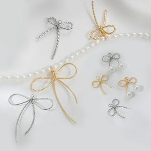 May include: An assortment of delicate bow-shaped charms in silver and gold tones. Some charms feature dangling faux pearls. The charms are arranged on a white surface with a strand of pearls, suggesting jewelry-making supplies or decorative elements.