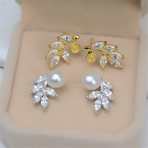 May include: A pair of earrings featuring a pearl and a leaf design. The earrings are made of silver and have a gold finish. The leaves are decorated with clear cubic zirconia stones.