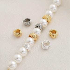 Può includere: Una composizione di perline e accessori per gioielli. L'immagine presenta un filo di grandi perle bianche, simili a perle. Ad accompagnare il filo ci sono diverse piccole perline decorative avvolte in filo metallico in tonalità oro e argento.